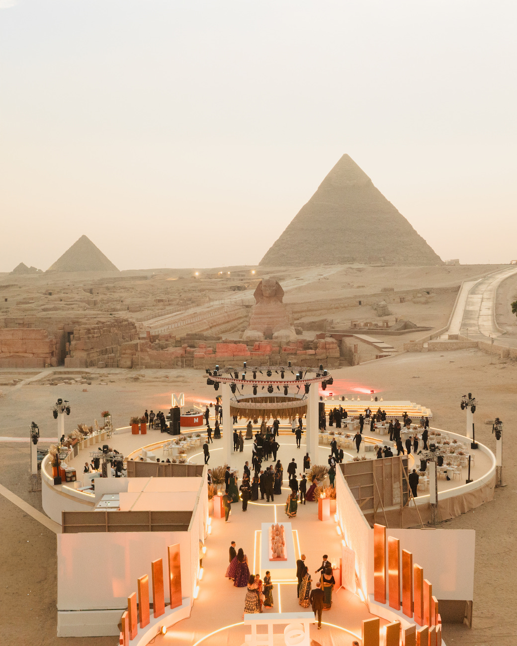 People walk on a decorated pathway toward an event space with the Pyramids of Giza visible in the background under a hazy sky.
