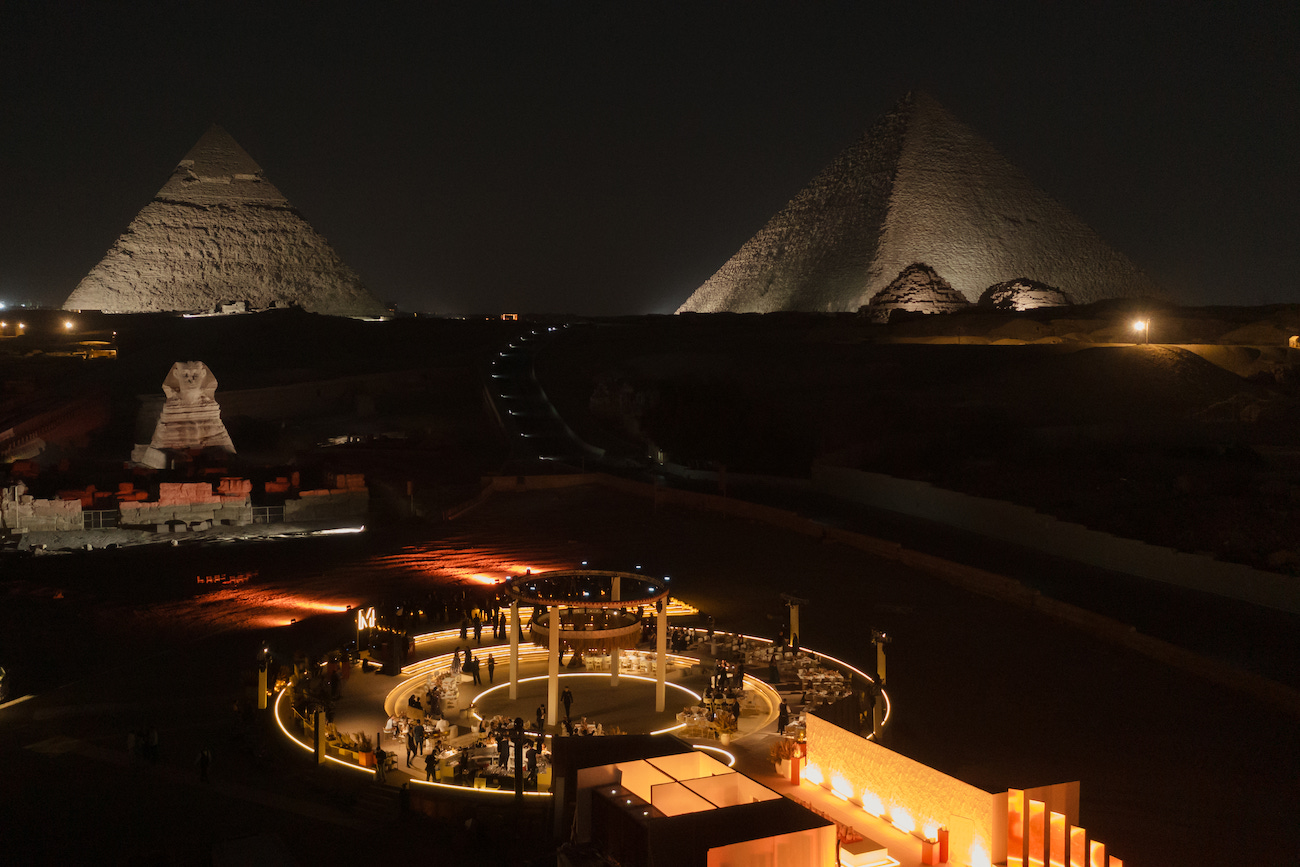 The Pyramids of Giza and the Sphinx are illuminated at night, with spotlights highlighting their structures and a lit circular area in the foreground.