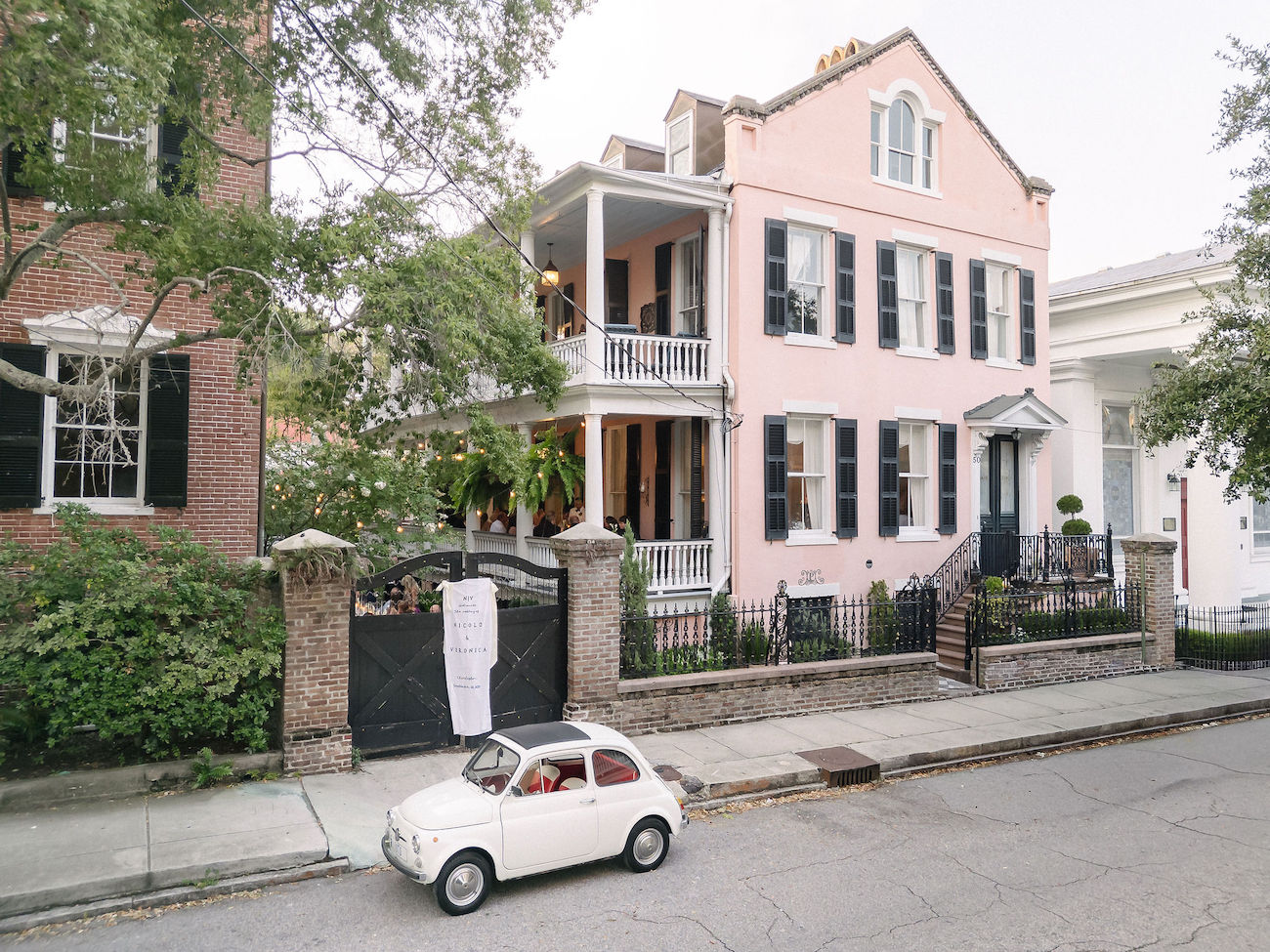 A small white vintage car is parked on the street in front of a pink two-story house with black shutters and a wraparound balcony.