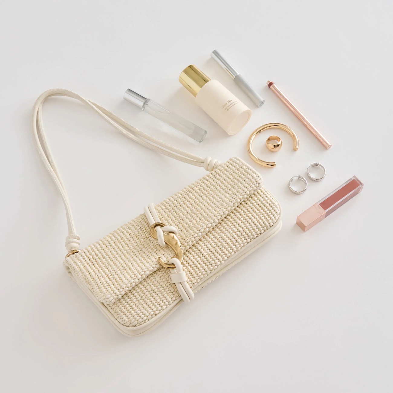 A woven cream handbag with a gold clasp is placed next to rings, lip gloss, a nail polish bottle, a face mist, a compact, and a bracelet on a white surface.