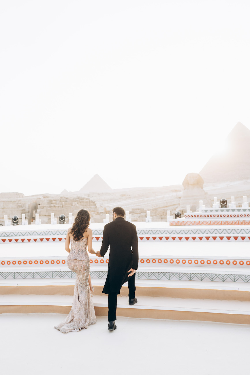 A couple dressed in formal attire walks up steps toward an outdoor area with pyramids visible in the background under bright sunlight.