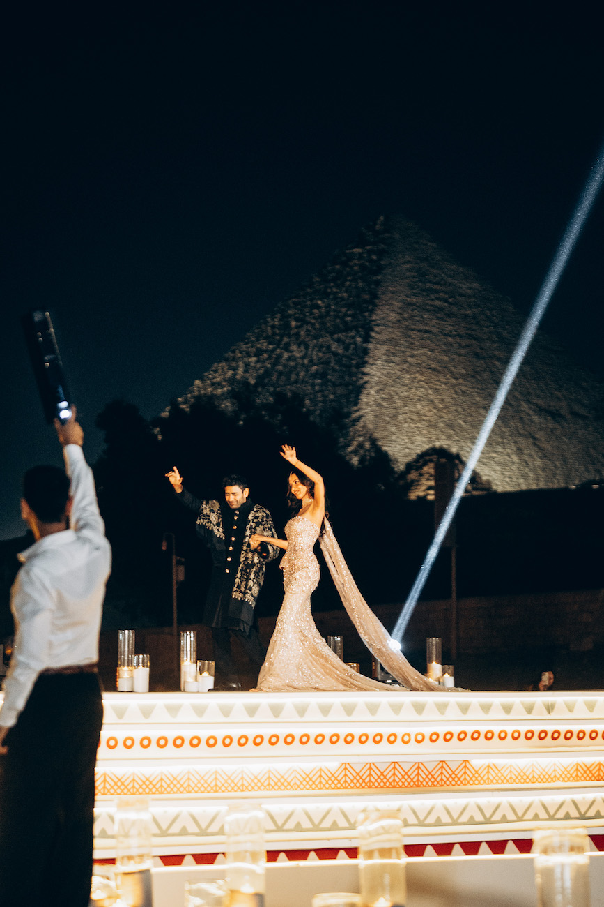 A bride and groom stand on a decorated platform at night, celebrating in front of an illuminated pyramid, with a person in the foreground raising an object.