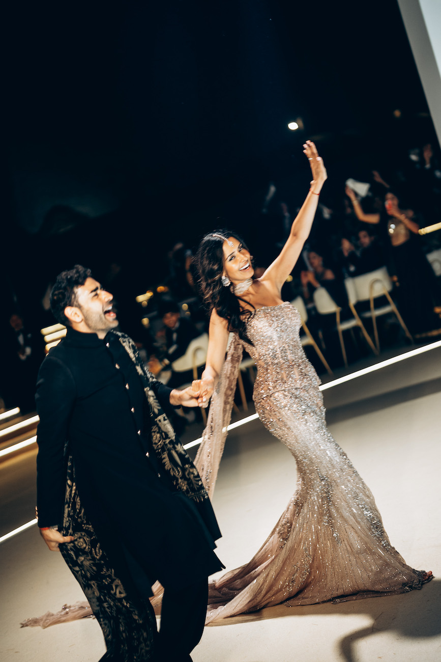 A woman in a sparkly gown waves while walking with a man in traditional attire at a formal event, with people seated in the background.