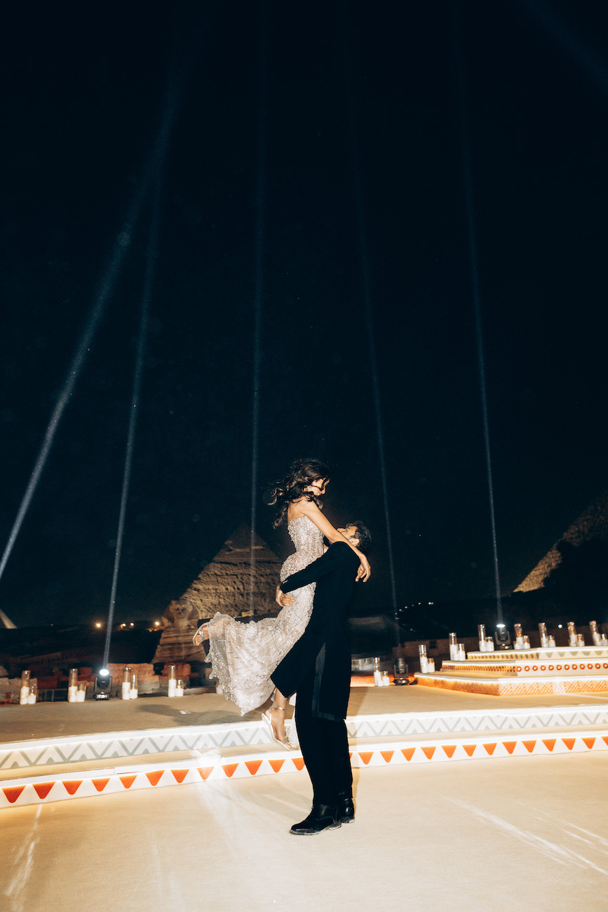 A person in formal attire lifts another person in a white dress at night, with pyramids and spotlights visible in the background.