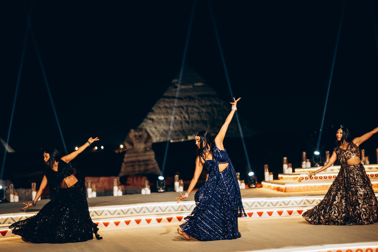 Three women in traditional attire perform a synchronized dance outdoors at night, with a large, illuminated pyramid structure in the background.