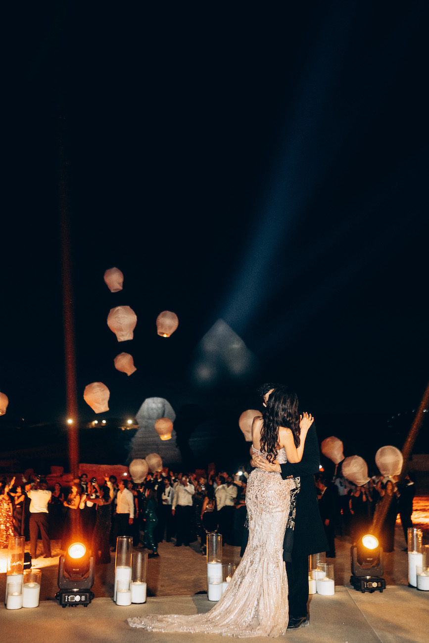 A couple dances at night in front of a crowd, with paper lanterns floating in the sky and the pyramids illuminated in the background.