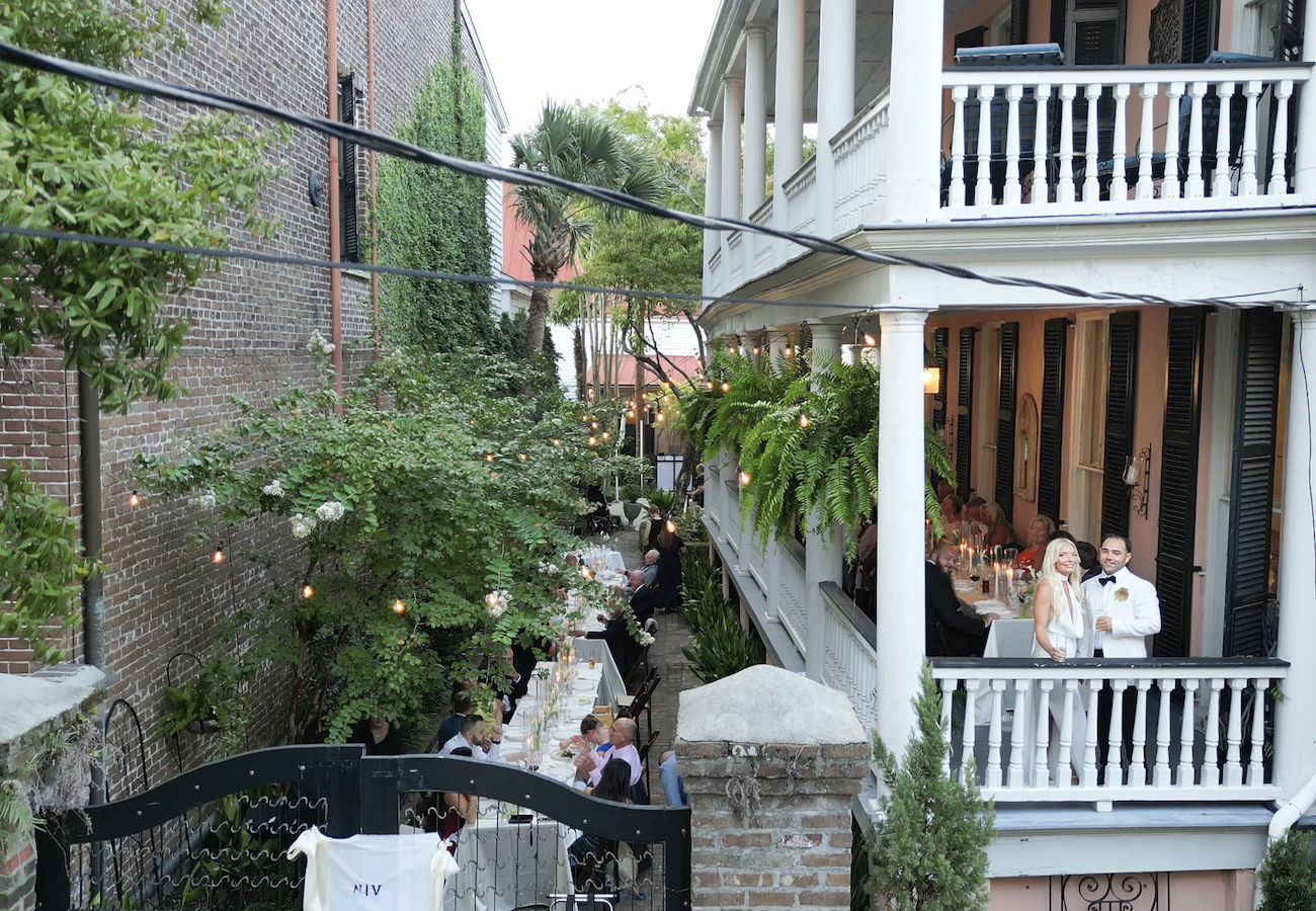 Outdoor dining setup in a narrow courtyard next to a brick building, with tables arranged under string lights; people gathered on both the patio and upper balcony.