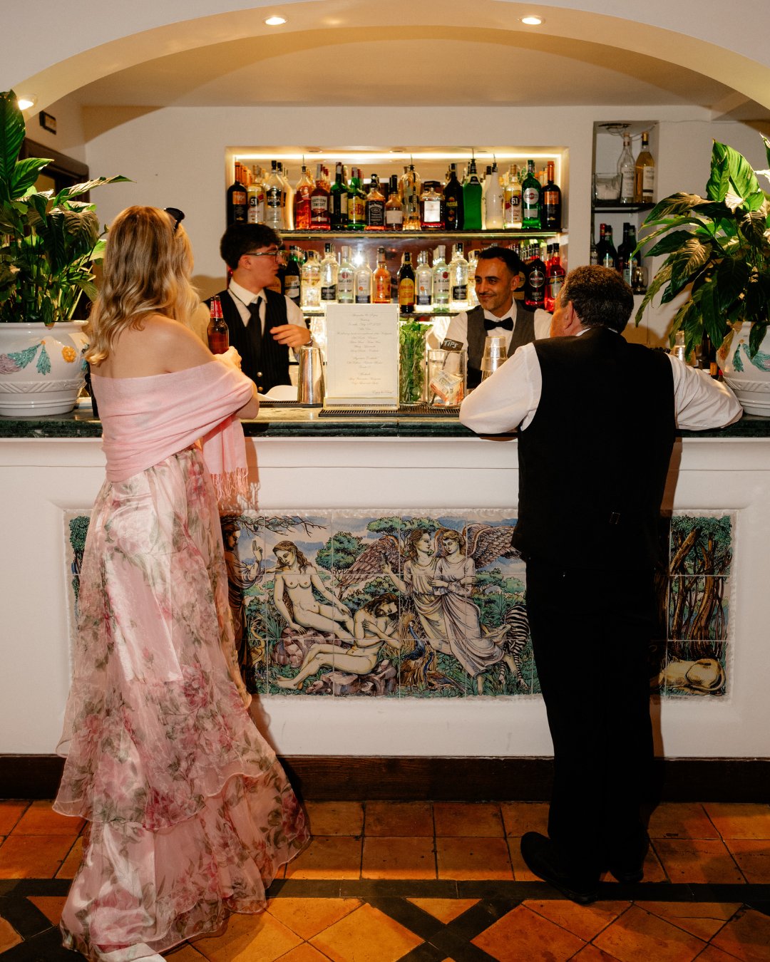 A woman in a pink floral dress and two men in formal attire stand at a bar with bottles and plants in the background.