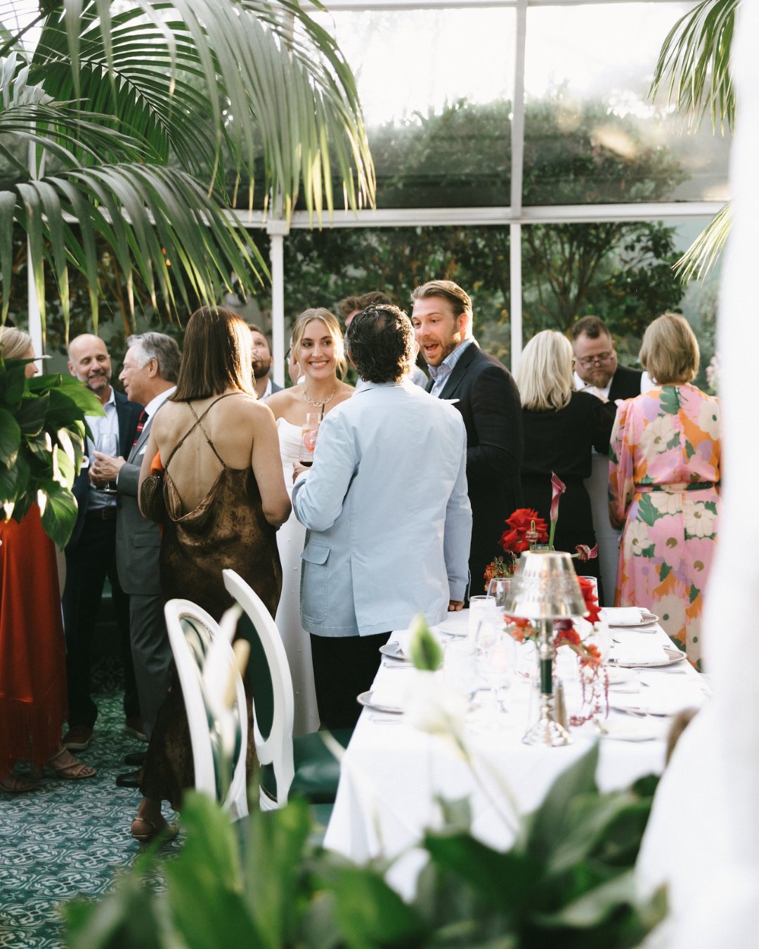 A group of people dressed in formal attire are socializing in a bright, plant-filled room near a dining table set for an event.