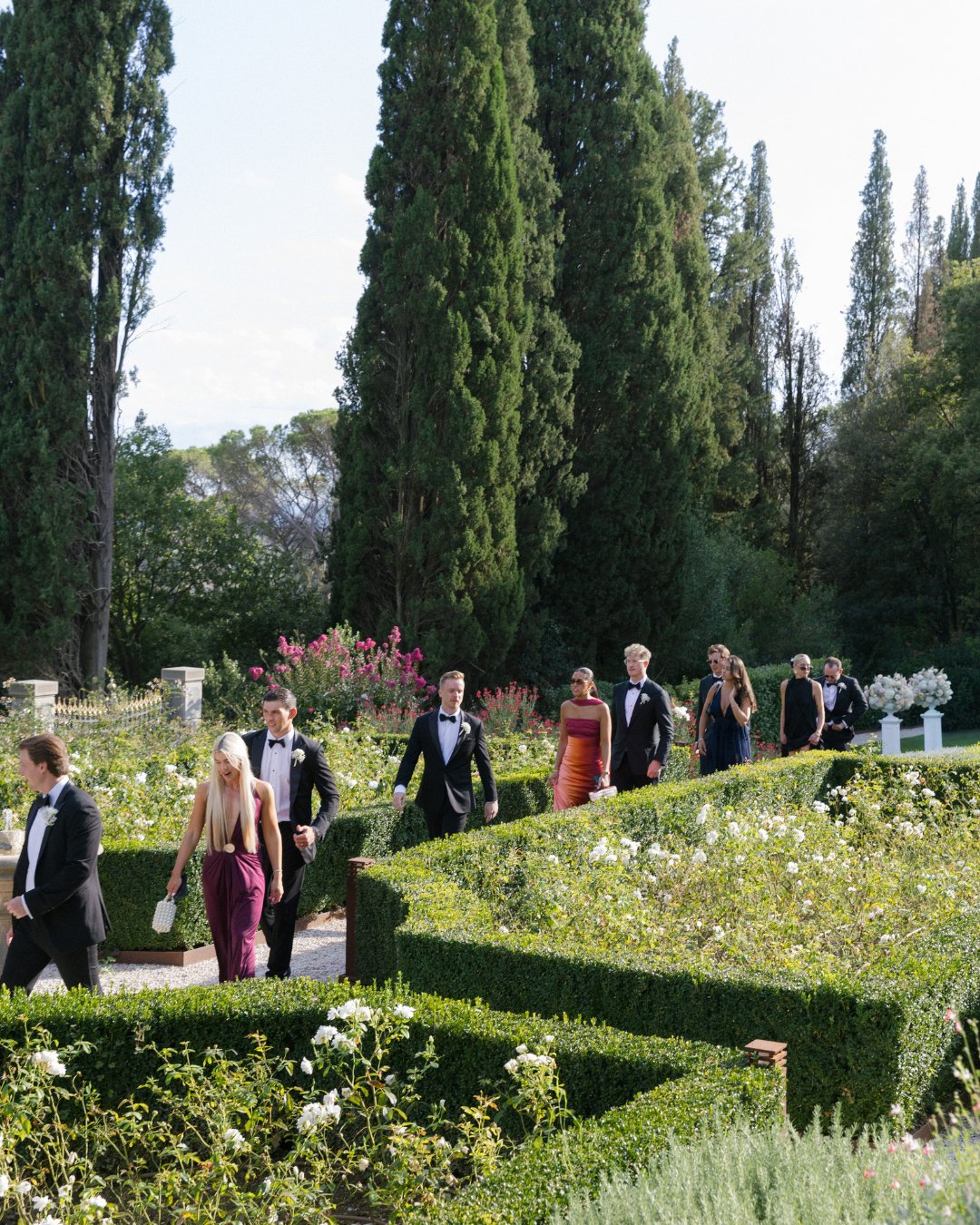 A group of people in formal attire walk along a garden path bordered by manicured hedges and tall trees.