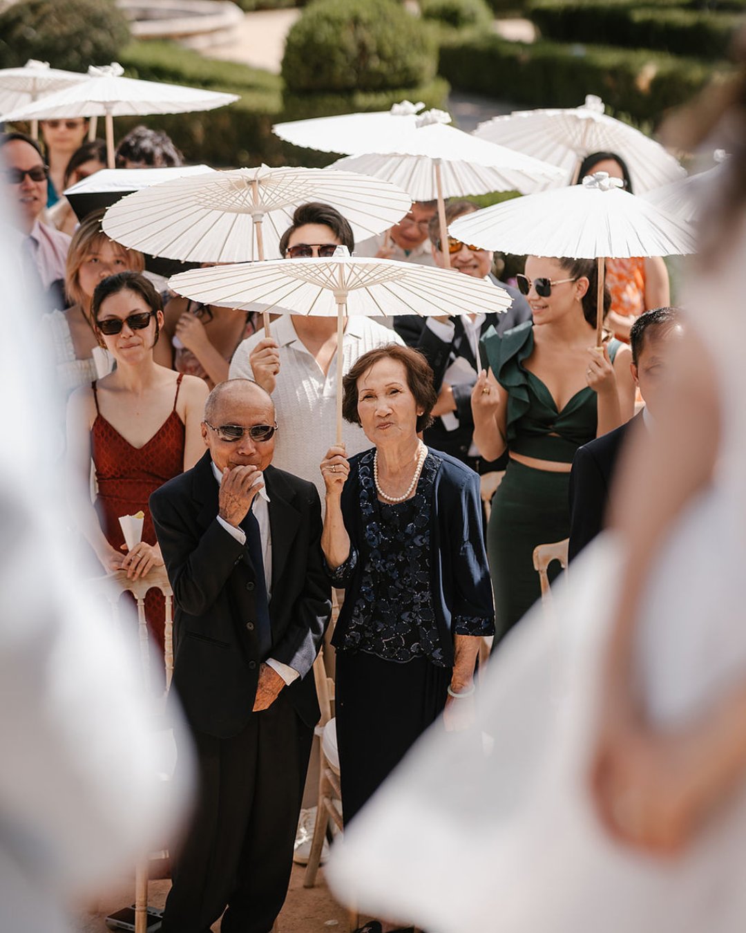 A group of people, some holding white parasols, stand outdoors at a formal event, with two individuals in focus looking forward.