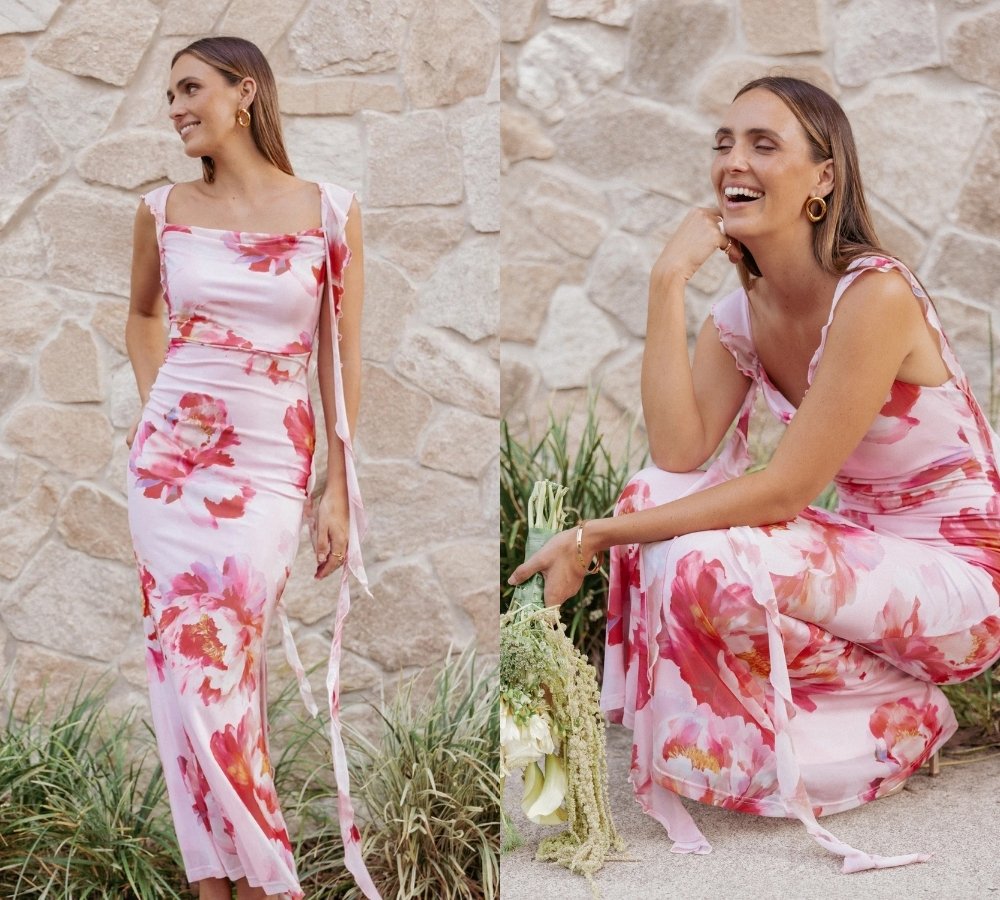 A woman wearing a long, sleeveless pink dress with red floral prints stands and sits in front of a stone wall, smiling and holding a bouquet of flowers.