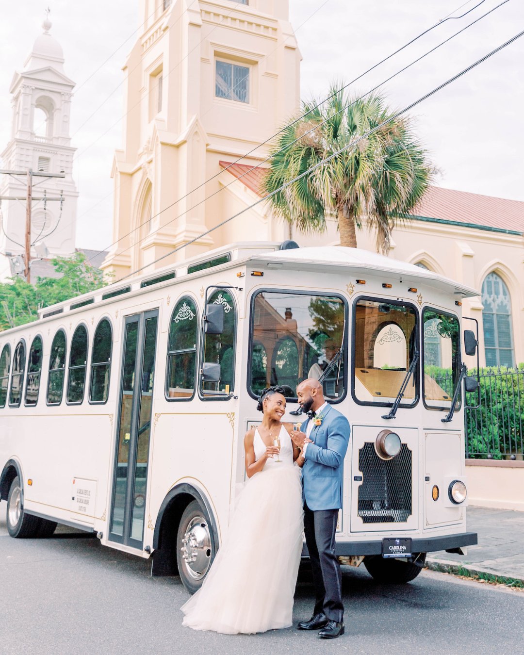 A bride and groom stand together in front of a white trolley parked on a street with a church in the background.