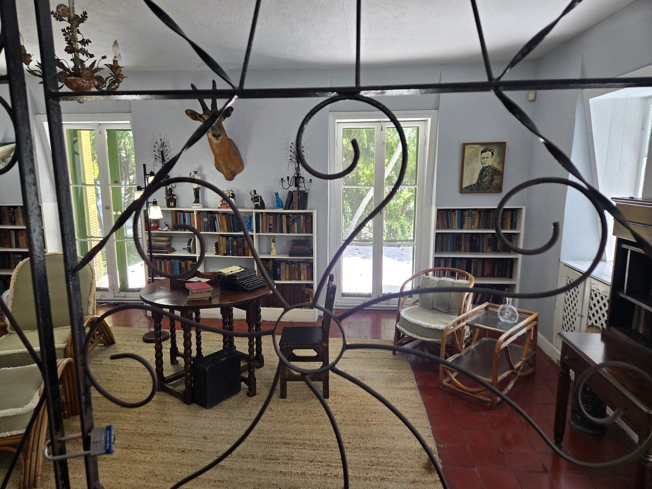 A view of the Ernest Hemingway home living space through an ornate iron gate into a sunlit room with bookshelves, armchairs, a small table, and various decorative items.