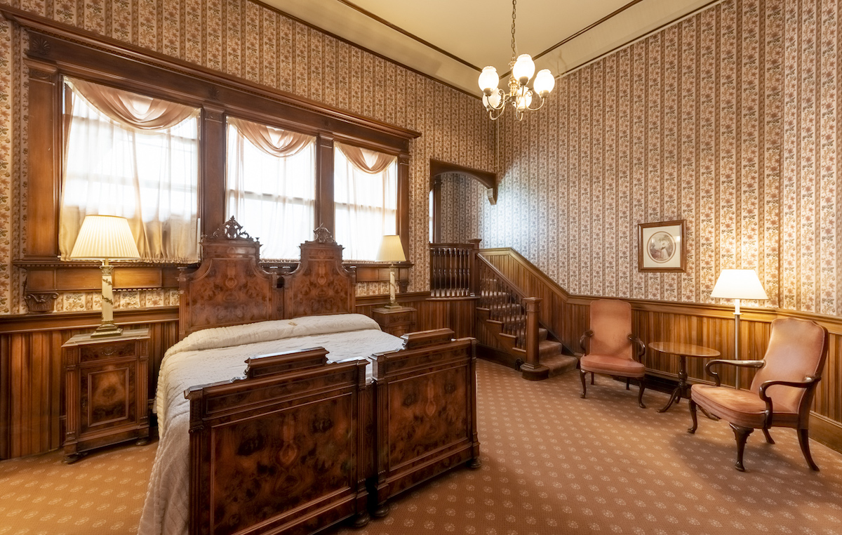 Victorian-style bedroom with ornate wood furniture, patterned wallpaper, two armchairs, lamps, and a staircase leading to an upper level.