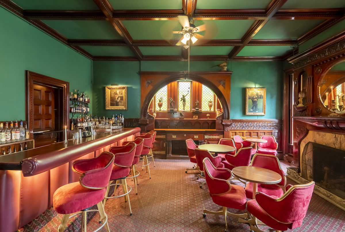 A vintage-style bar with red velvet chairs, green walls, a wooden ceiling, a fireplace, and a curved archway leading to another room.