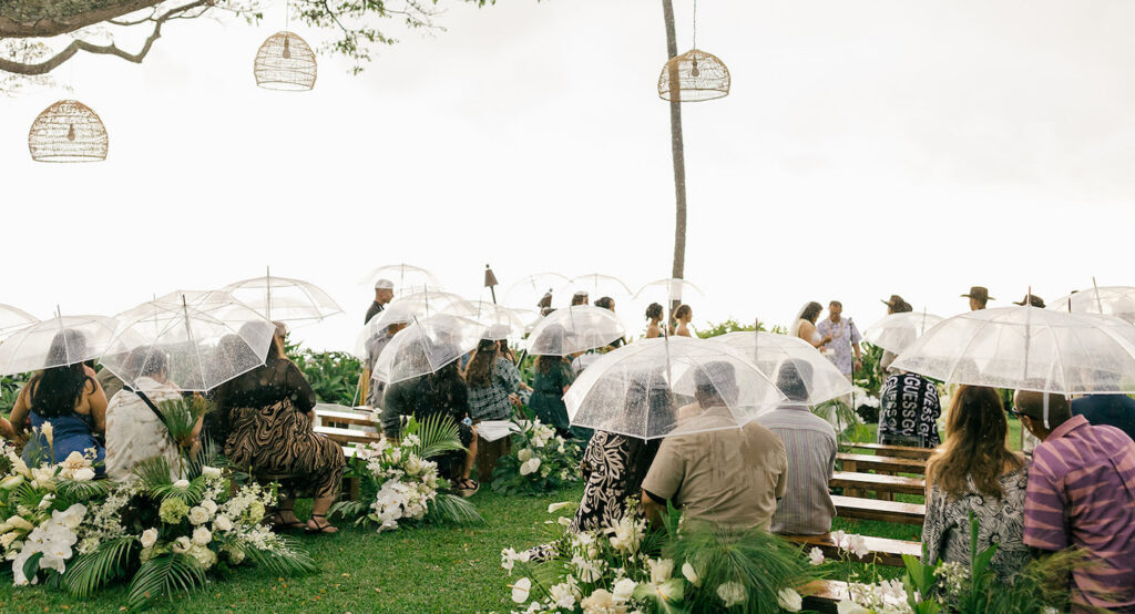 Guests sit on benches holding clear umbrellas at an outdoor wedding ceremony on a grassy lawn, surrounded by flowers and greenery.