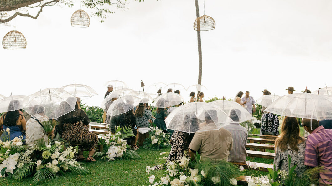 Guests sit on benches holding clear umbrellas at an outdoor wedding ceremony on a grassy lawn, surrounded by flowers and greenery.