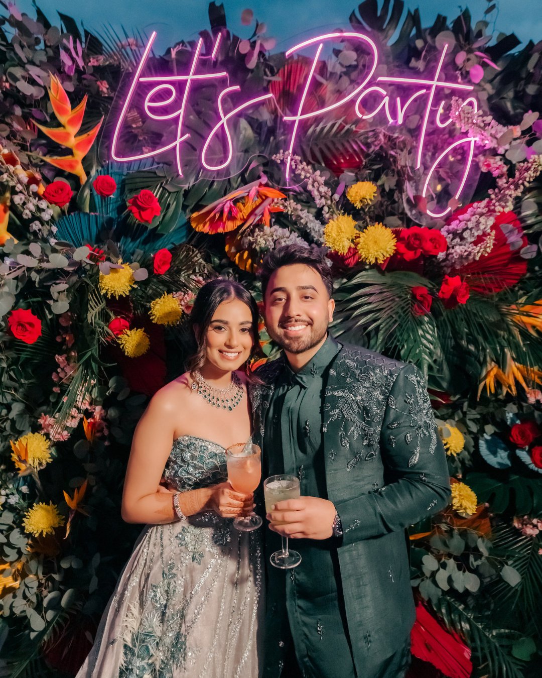 A couple dressed in formal attire holds drinks and smiles in front of a colorful floral backdrop with a neon sign that reads "Let's Party."