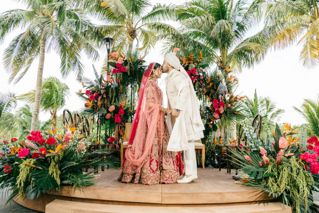 A bride and groom in traditional attire kiss on a decorated outdoor stage surrounded by tropical plants and colorful flowers.
