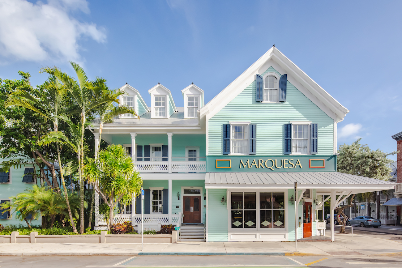A two-story pastel green building with white trim and blue shutters, labeled "Marquesa," stands on a sunny street corner with palm trees nearby.