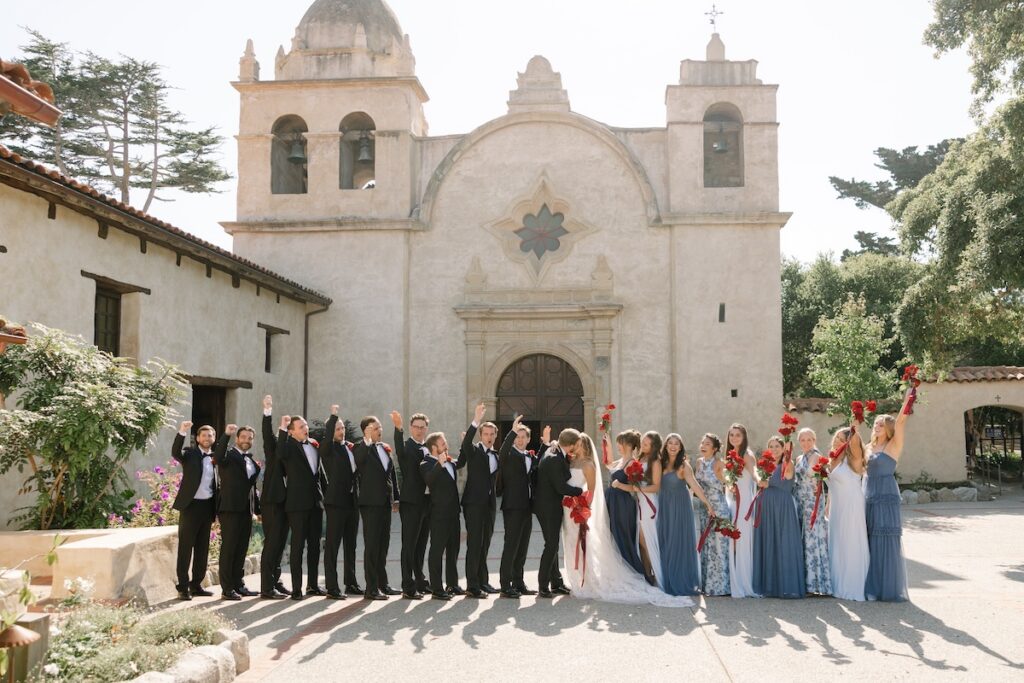 A wedding party poses in front of a historic church, with bridesmaids in blue dresses and groomsmen in black suits holding red bouquets and raising their hands.