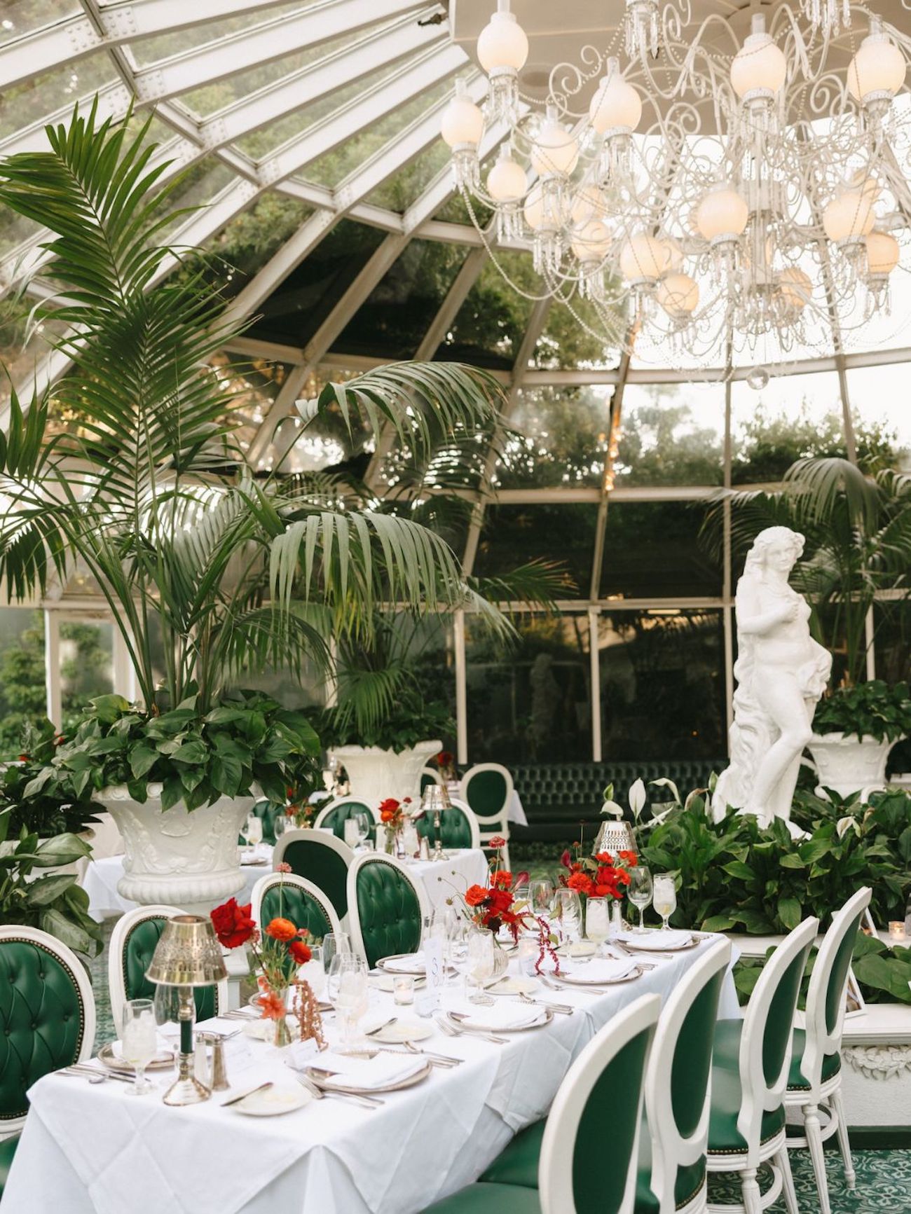 Elegant restaurant with green chairs, white-clothed tables set for dining, large indoor plants, a white statue, and a glass ceiling with a chandelier.