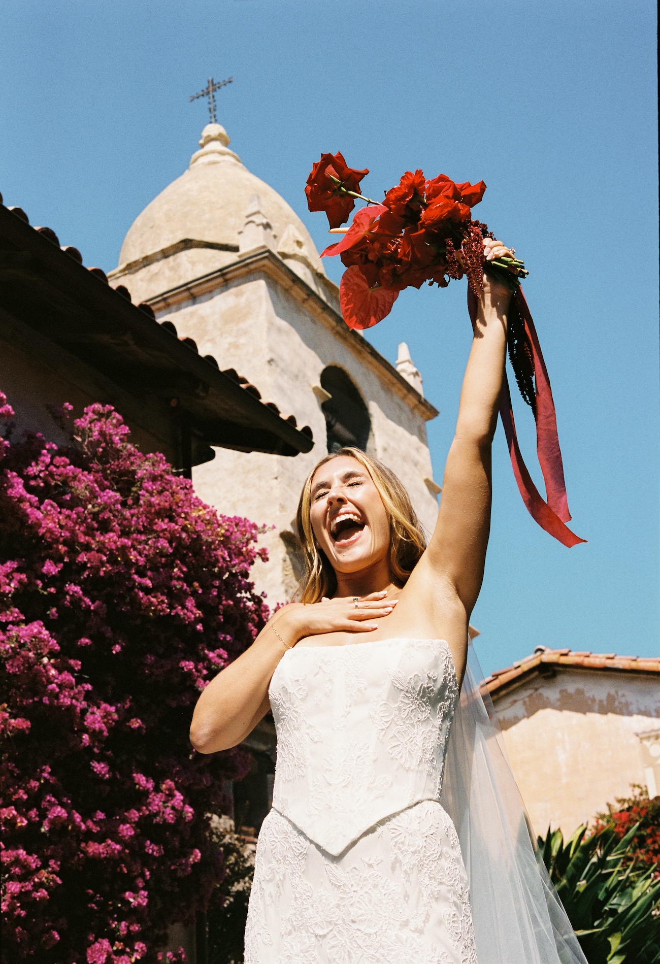 A bride in a white dress holds a bouquet aloft and smiles joyfully outside a church with a bell tower and blooming flowers.