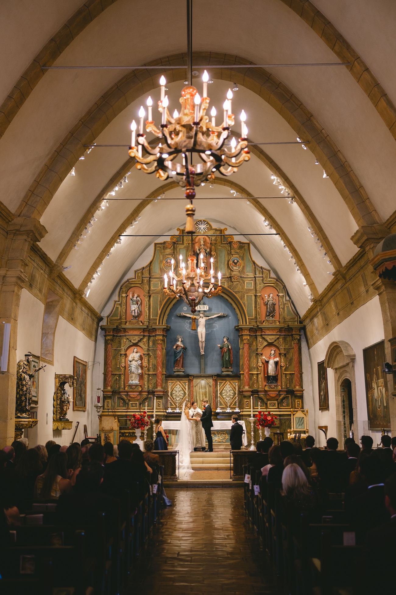 A wedding ceremony is taking place at the altar of a church, with guests seated in pews and a chandelier hanging from the arched ceiling.