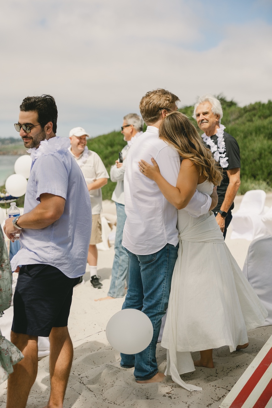 A bride and groom hug barefoot on a sandy beach during a wedding ceremony, surrounded by casually dressed guests.