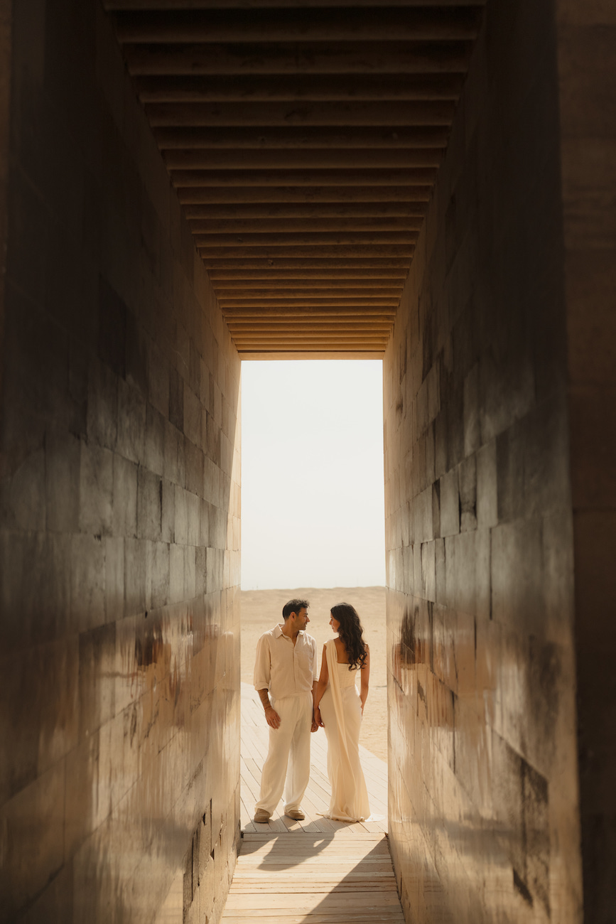 A couple stands facing each other at the end of a narrow corridor with sunlight and a beach visible in the background.