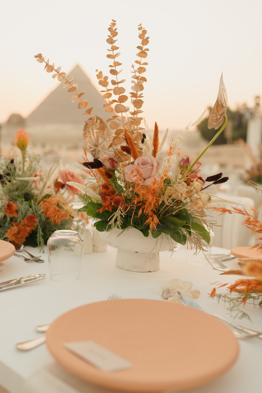A table is set with peach-colored plates, cutlery, and a centerpiece of dried and fresh flowers, with a pyramid visible in the background.