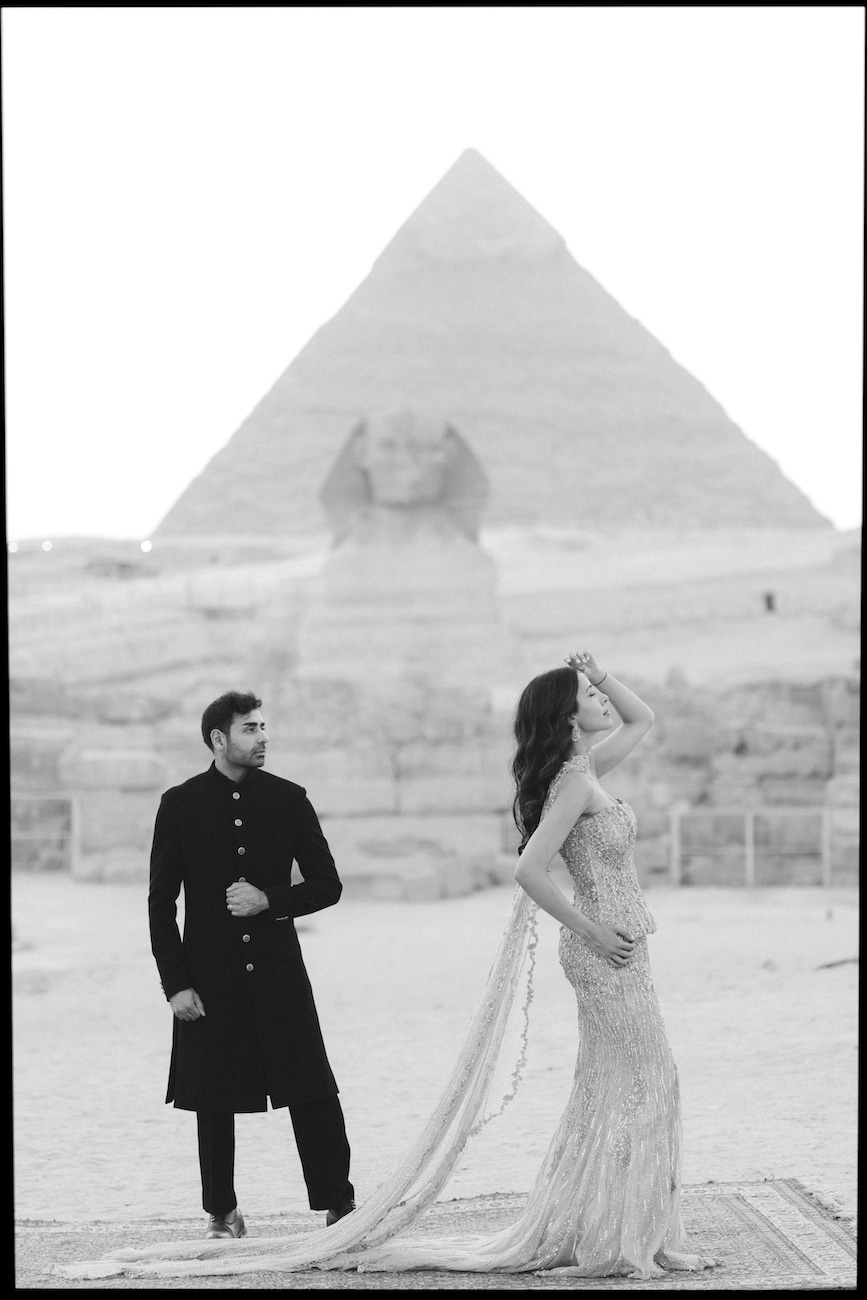 A man and woman pose in formal attire in front of the Great Sphinx and a pyramid in Egypt. The man looks at the woman while she gazes away, touching her hair.