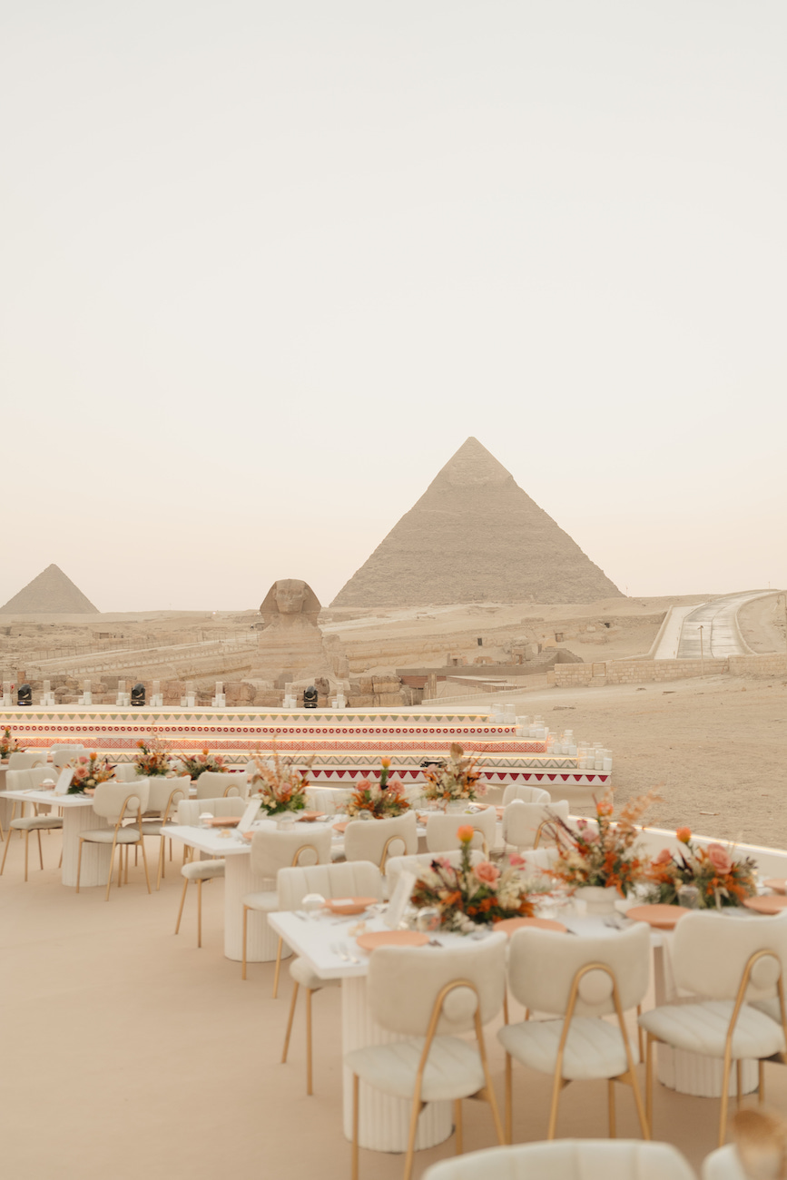 Elegant outdoor dining setup with decorated tables and chairs in front of the Pyramids of Giza and the Sphinx at sunset.