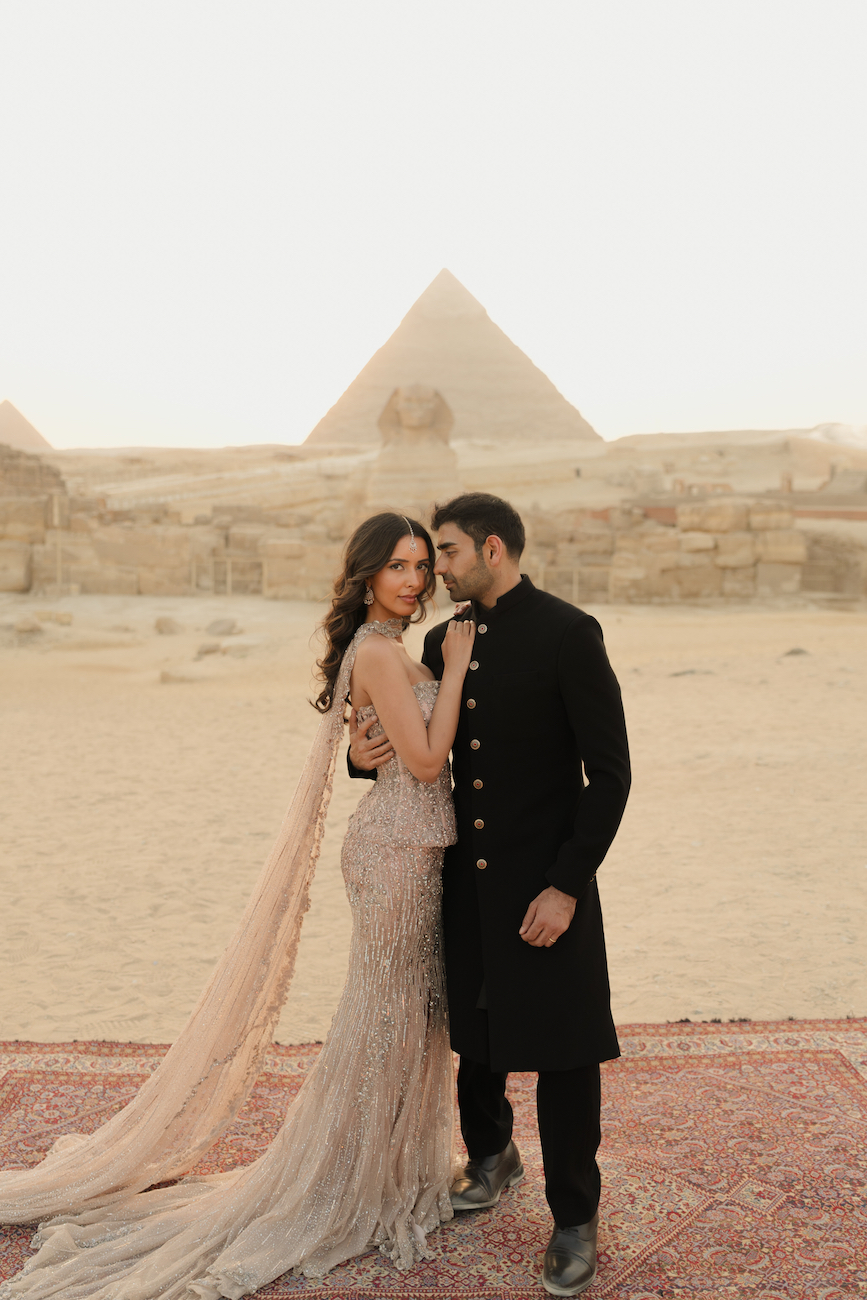 A couple poses together on a rug in front of the Pyramids of Giza; the woman wears a gown with a train, and the man wears a black traditional outfit.