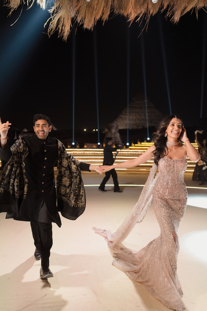 A man in a black traditional outfit and a woman in a glittering gown hold hands and walk on a stage with spotlights and a decorated background.