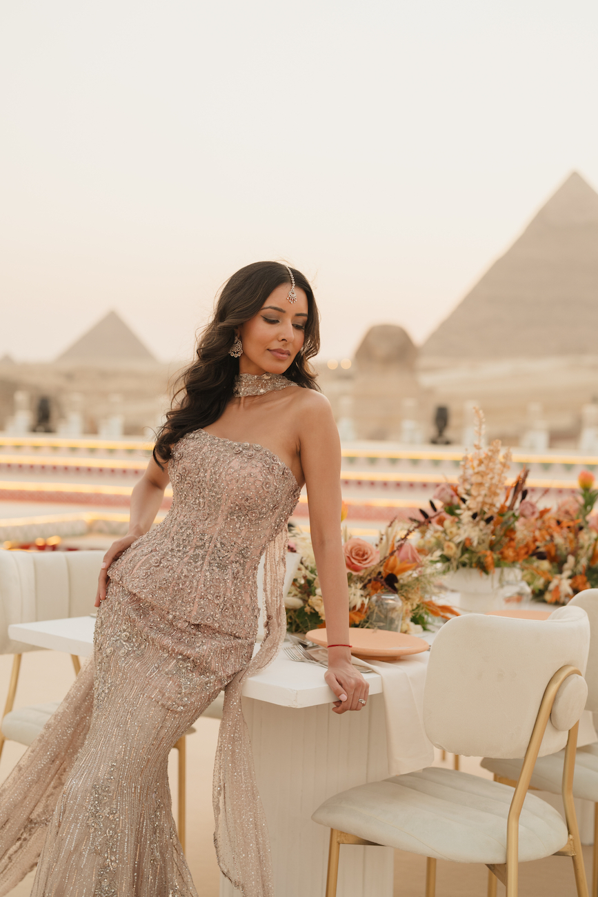 A woman in a strapless, sequined gown poses beside a decorated table with flowers, with the pyramids visible in the background at sunset.