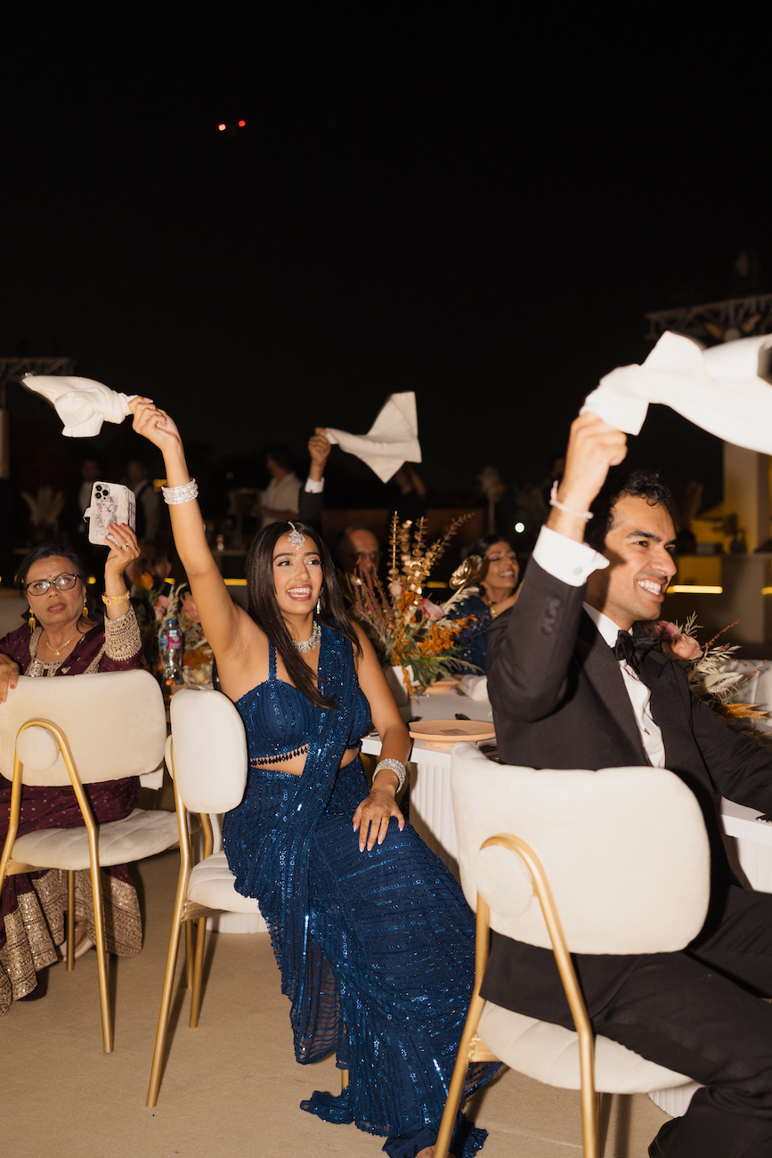 Guests seated at a table wave white napkins in the air during an evening event; one woman wears a blue dress, and a man beside her is in a suit.