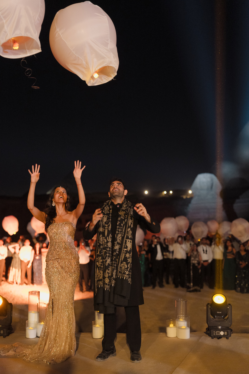 A woman in a gold dress and a man in black release sky lanterns at night; a crowd watches in the background.