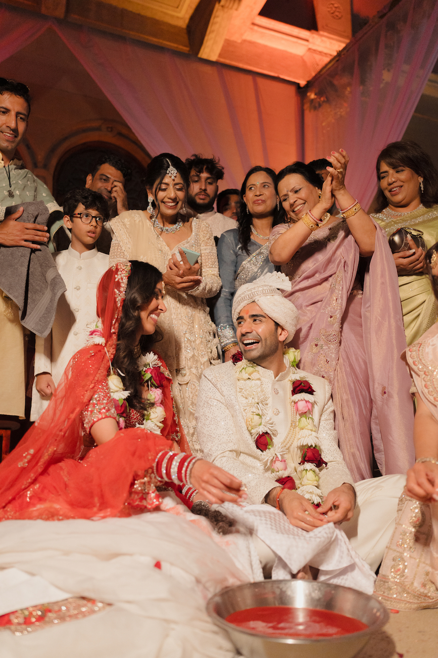 A bride and groom in traditional attire sit surrounded by smiling family and friends during a wedding ceremony.