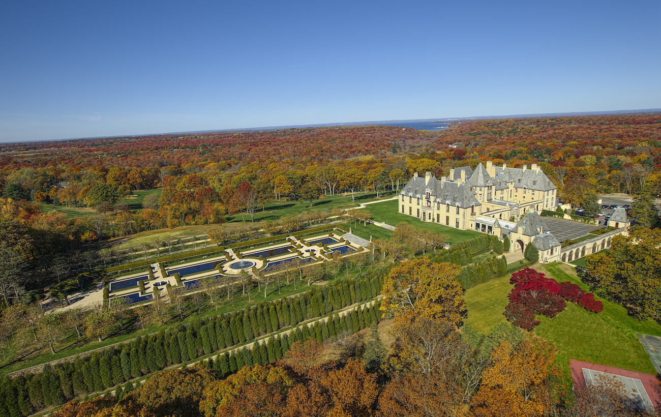 Aerial view of a large mansion with formal gardens, surrounded by autumn trees and extensive grounds under a clear blue sky.