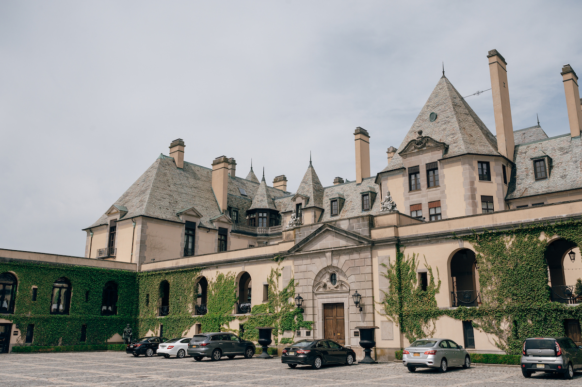 Large historic mansion with gray stone walls, multiple peaked roofs, and ivy-covered sections, with several cars parked in front on a paved lot.