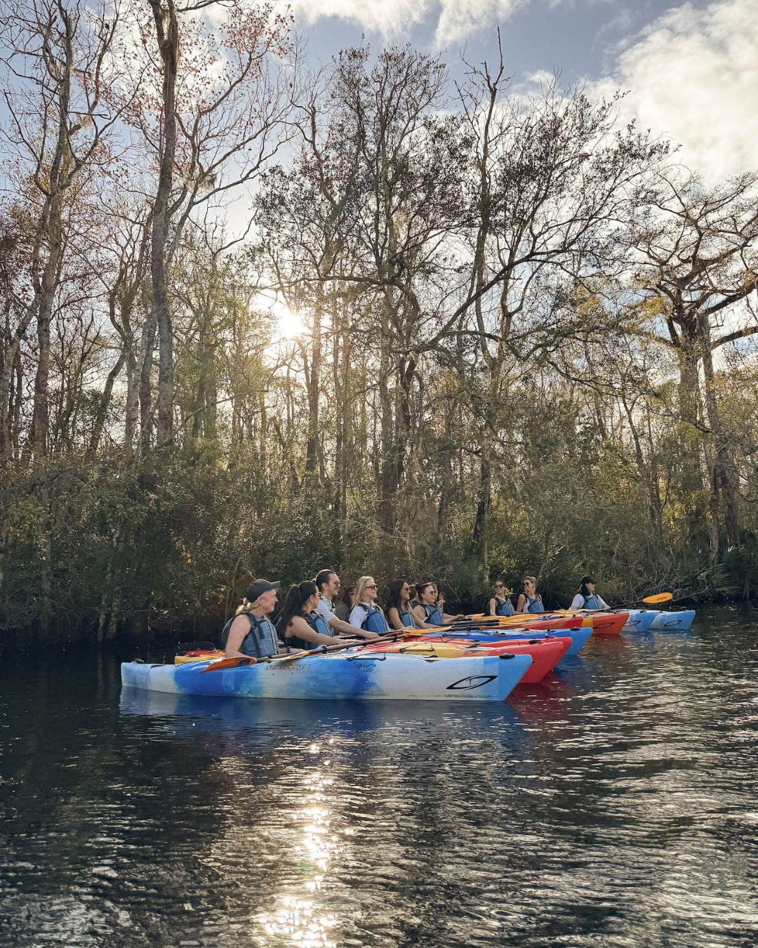 A group of people in colorful kayaks paddling on a calm river, surrounded by trees under a partly cloudy sky.