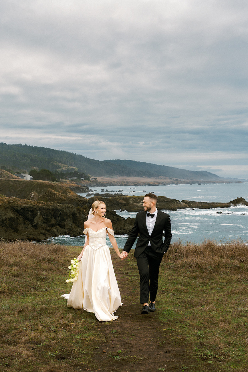 A bride and groom walk hand in hand outdoors near a rocky shoreline, with the ocean and hills in the background under a cloudy sky.