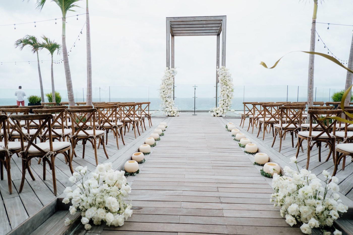 Outdoor wedding ceremony setup on a wooden deck with rows of chairs, white floral arrangements, and an arch overlooking the ocean.