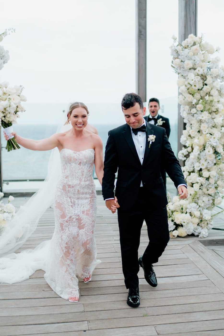 A bride in a lace wedding dress and a groom in a black tuxedo walk hand in hand on a wooden deck, surrounded by white floral arrangements.