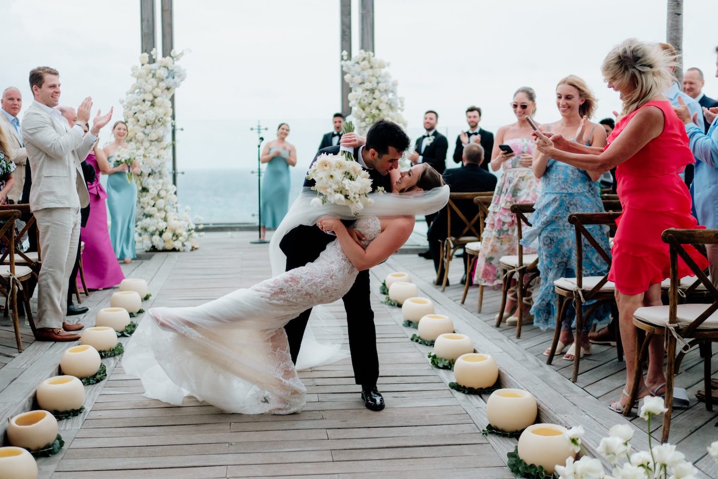 A bride and groom kiss as the groom dips the bride at an outdoor wedding ceremony, surrounded by applauding guests and floral decorations.