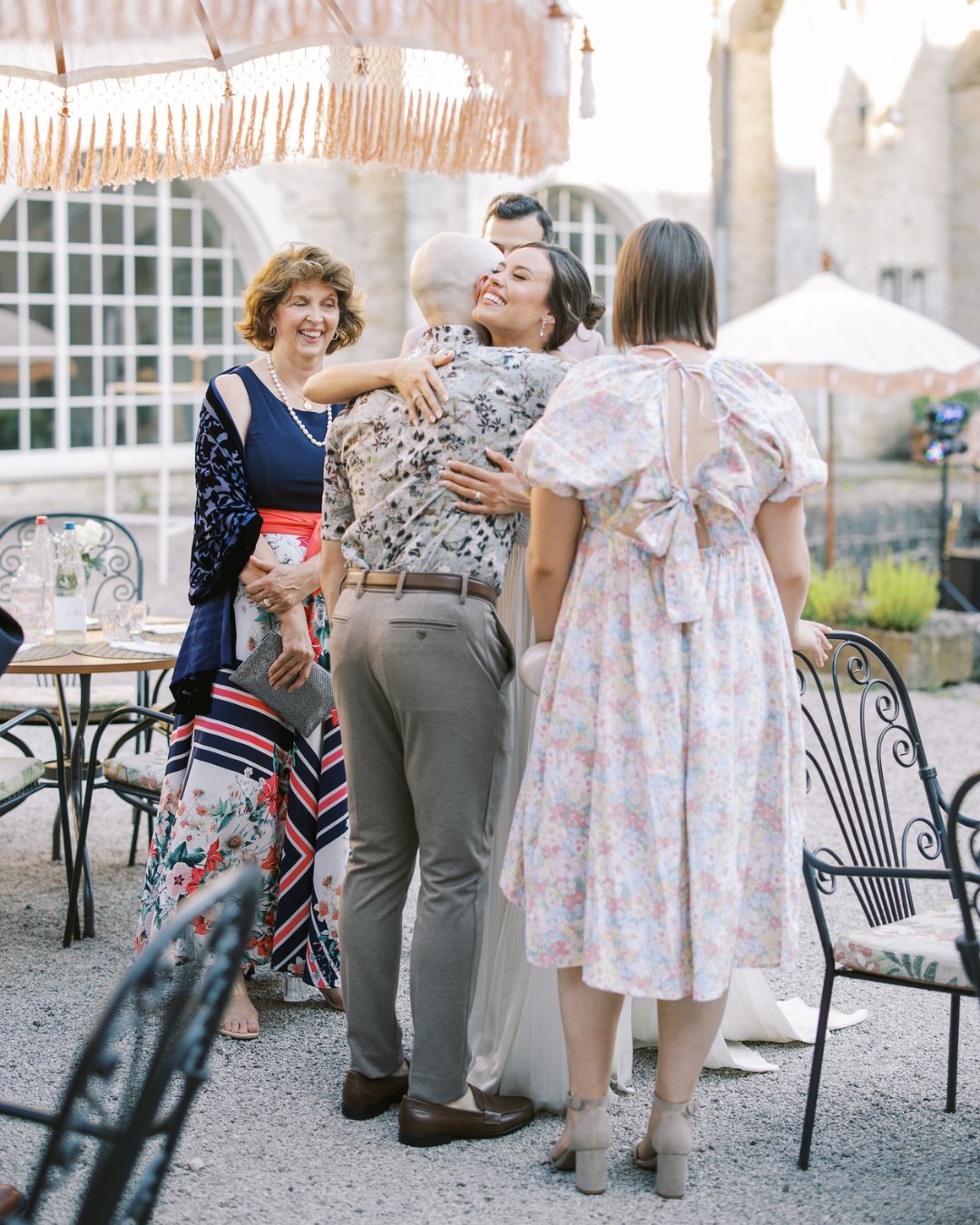 Four adults stand outside near a table; two hug while the other two watch and smile. The setting appears to be a courtyard with stone buildings and large windows.