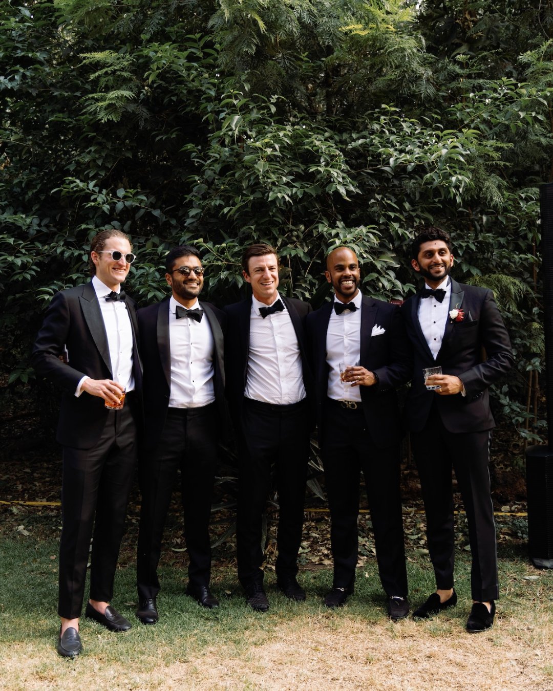 Five men dressed in black tuxedos stand together outdoors in front of greenery, smiling and holding drinks.