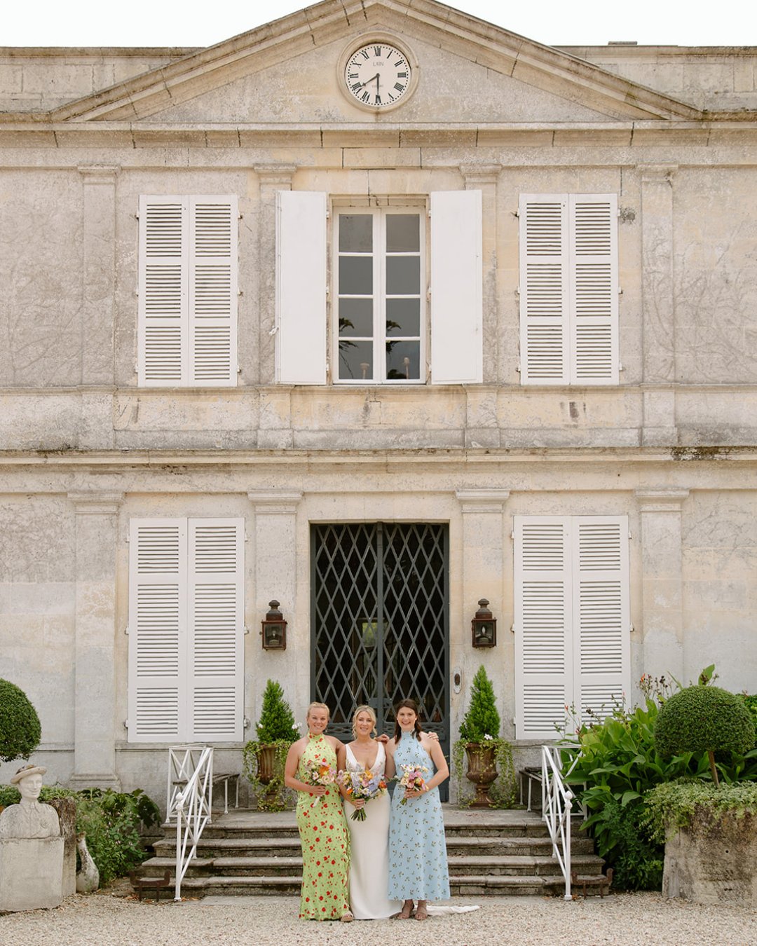 Three women in formal dresses stand holding bouquets in front of a large, light-colored building with white shutters and a clock above the entrance.